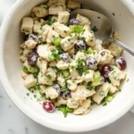 Bowl of chicken salad with diced chicken, halved red grapes, chopped celery, and fresh herbs, with a spoon resting inside the bowl; small bowls of extra chopped celery on the side.
